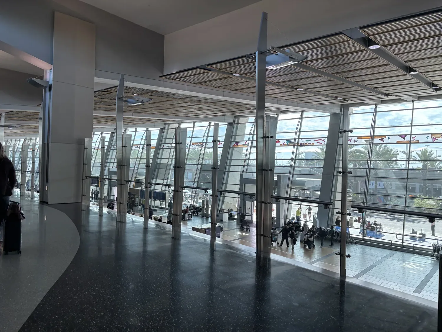 A spacious modern airport terminal interior with large glass windows, metal beams, and people walking or sitting