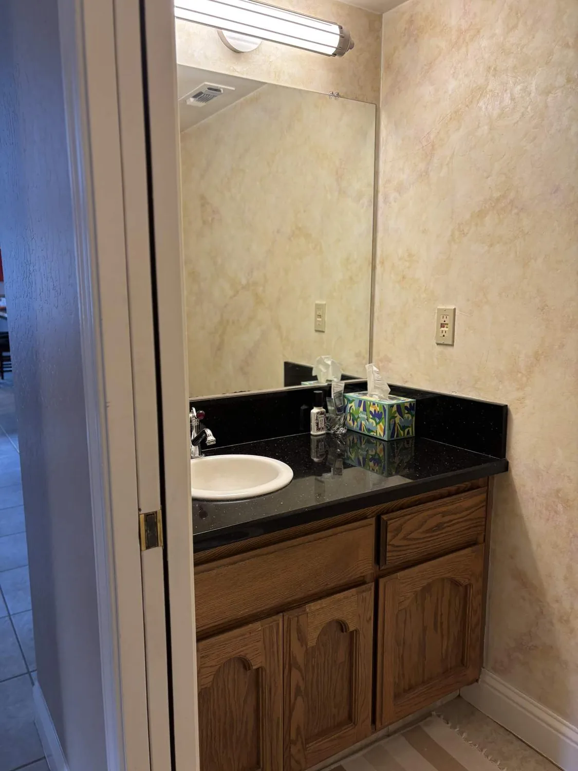 A bathroom vanity with a black countertop, wooden cabinets, a round white sink, and a large mirror above