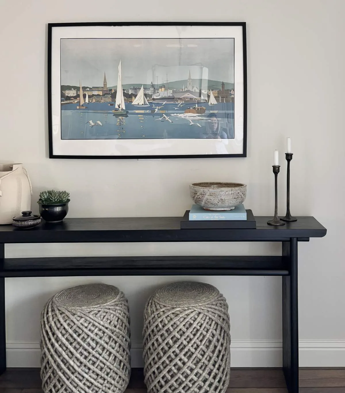 A minimalist styled entryway or living space with a black console table, decorative items, two woven stools, and a framed nautical-themed artwork on the wall