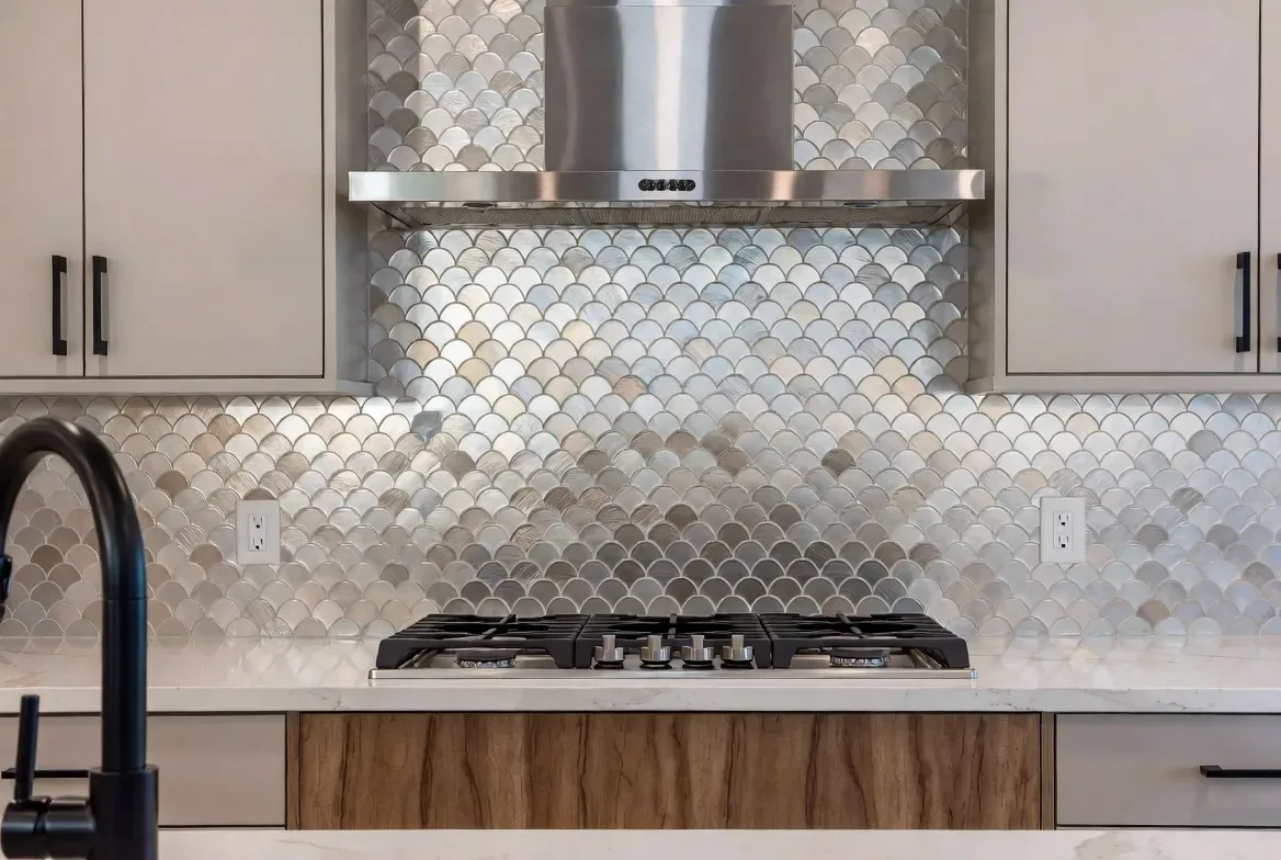 A modern kitchen featuring a metallic fish scale tile backsplash, stainless steel range hood, gas stove, and light cabinetry with black handles