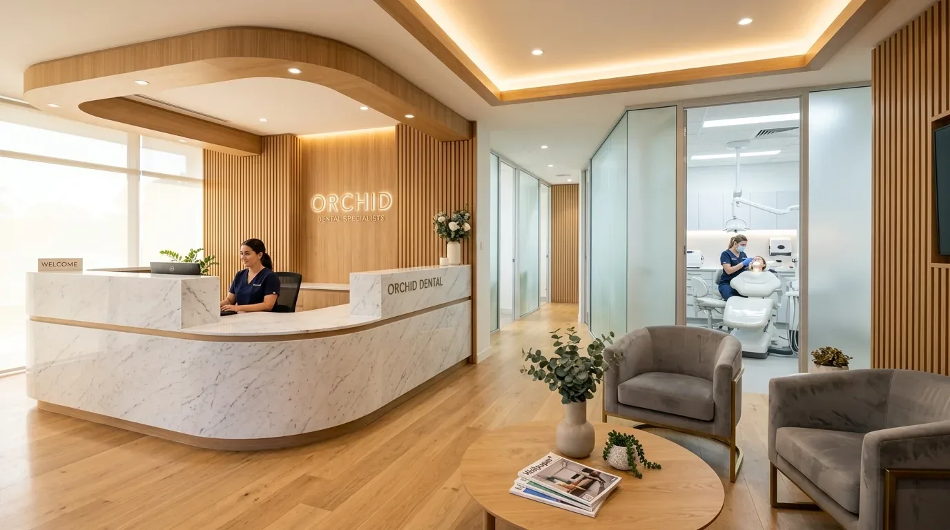 A modern dental clinic reception area with a marble desk, wooden accents, a waiting area with chairs and a table, and a visible treatment room where a dentist is attending a patient