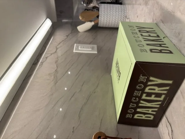A kitchen countertop with a Bouchon Bakery box, marble backsplash, under-cabinet lighting, and kitchen utensils in a holder