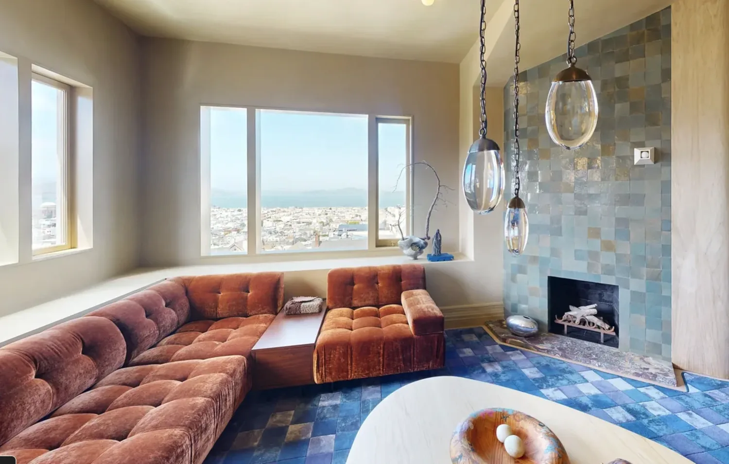 A modern living room with a large rust-colored sectional sofa, blue tiled fireplace, and large windows overlooking a cityscape