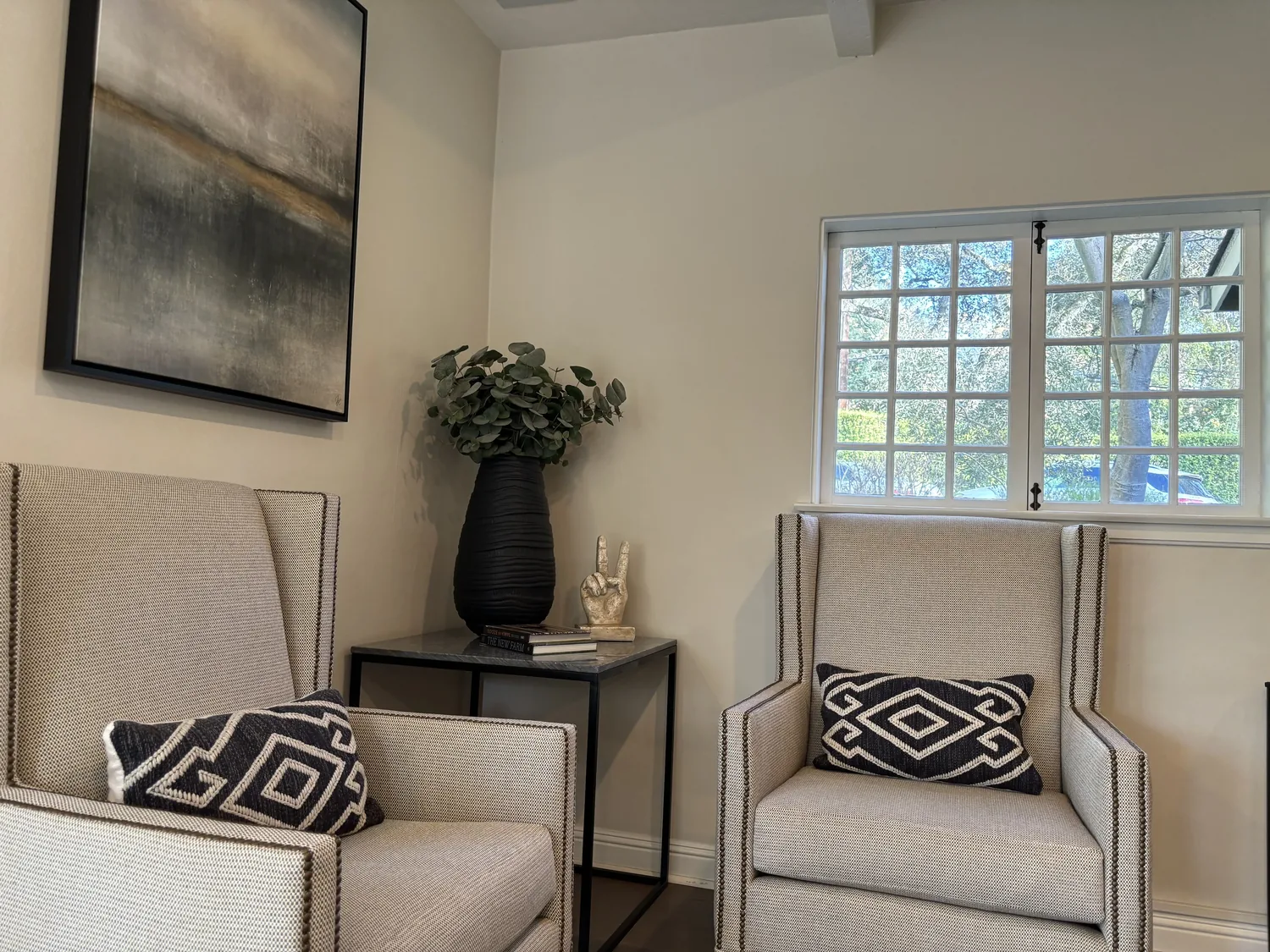 A cozy living room corner with two beige armchairs, a side table with books and a decorative hand sculpture, a large black vase with greenery, a window with grid panes, and an abstract painting on the wall