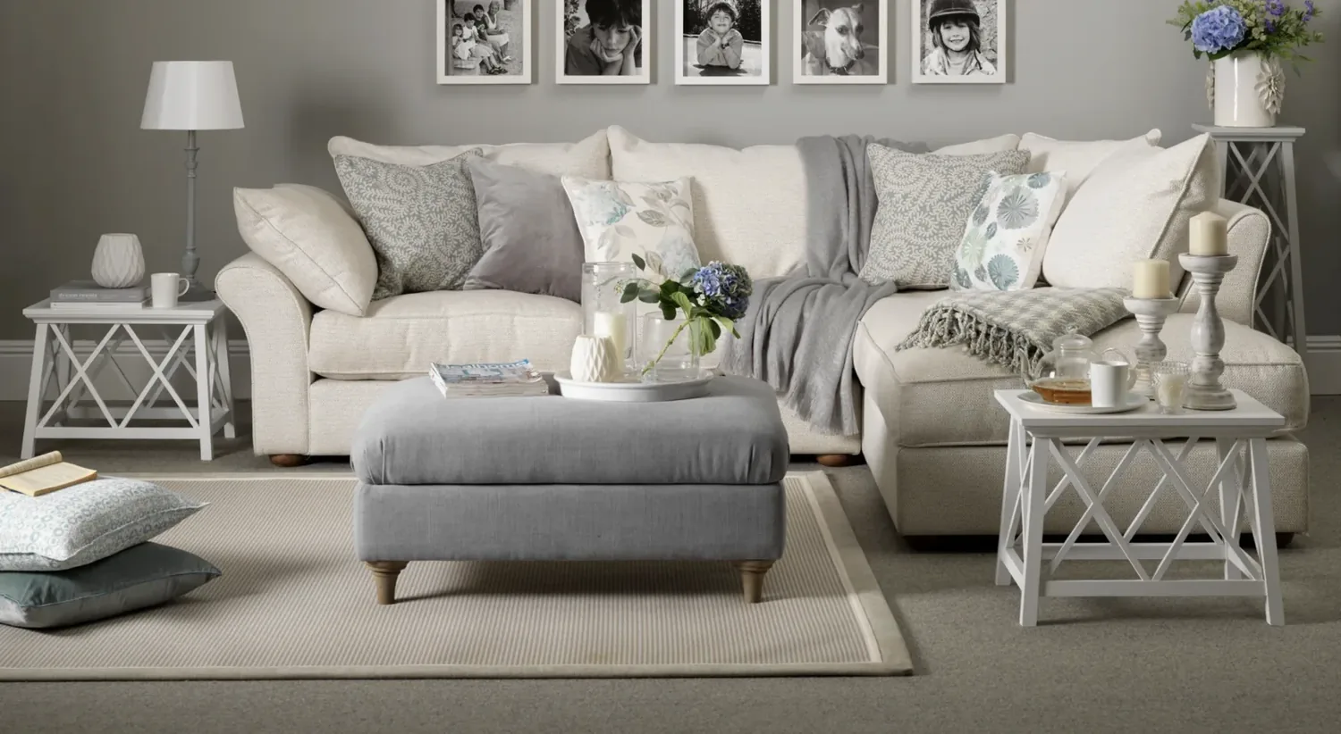 A cozy living room with a beige sectional sofa, gray ottoman, white side tables, decorative pillows, and framed black-and-white photos on the wall