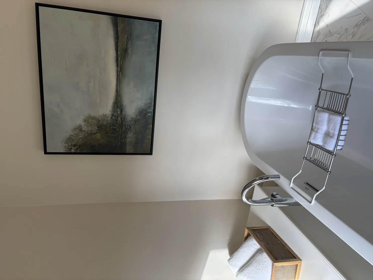 A modern bathroom featuring a white freestanding bathtub with a chrome faucet, a towel rack with white towels, and a large abstract painting on the wall