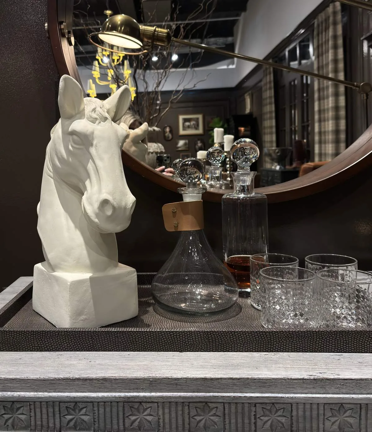A decorative tray with a white horse bust, glass decanters, and glasses in front of a round mirror in a dark-toned room